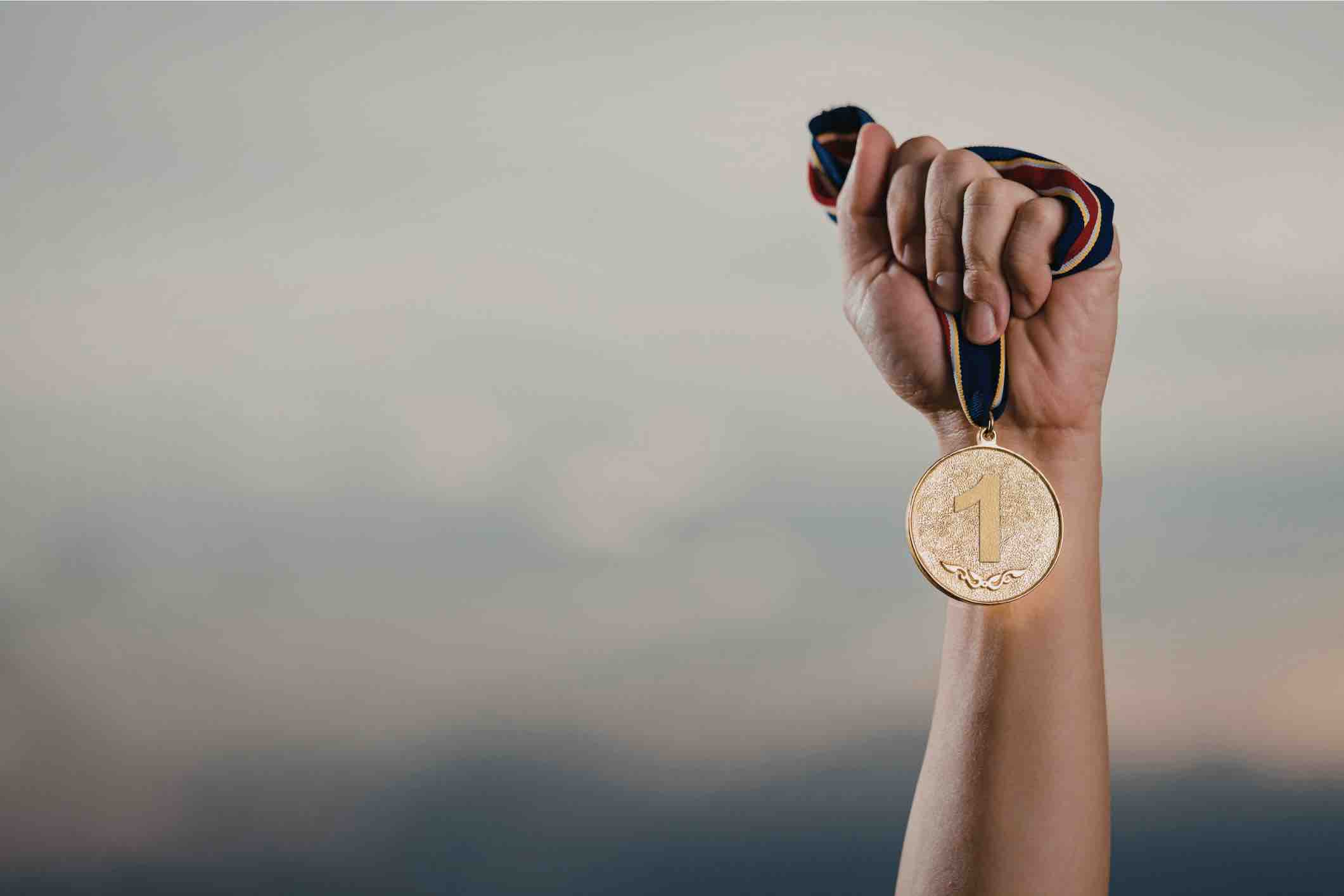 Hand holding a first-place gold medal against a blurred sky background.
